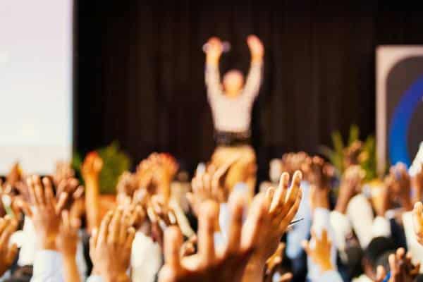 a room full of people are raising their hands and a speaker stands on a stage with his arms raised